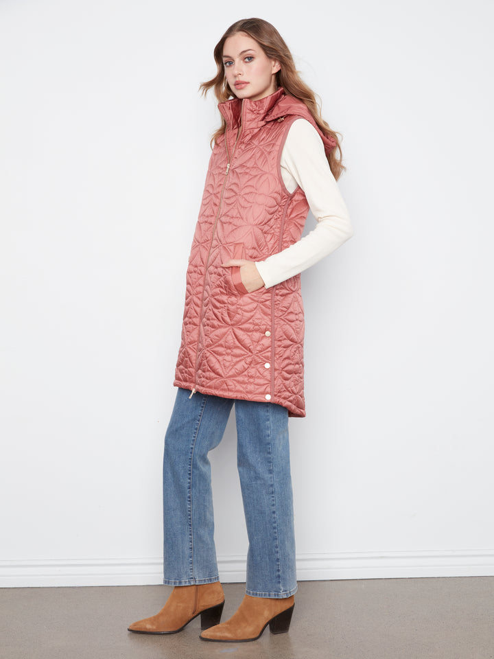 A woman modeling a dusty rose quilted sleeveless puffer vest over a cream long-sleeve top, blue jeans, and tan heeled ankle boots, standing against a white wall.