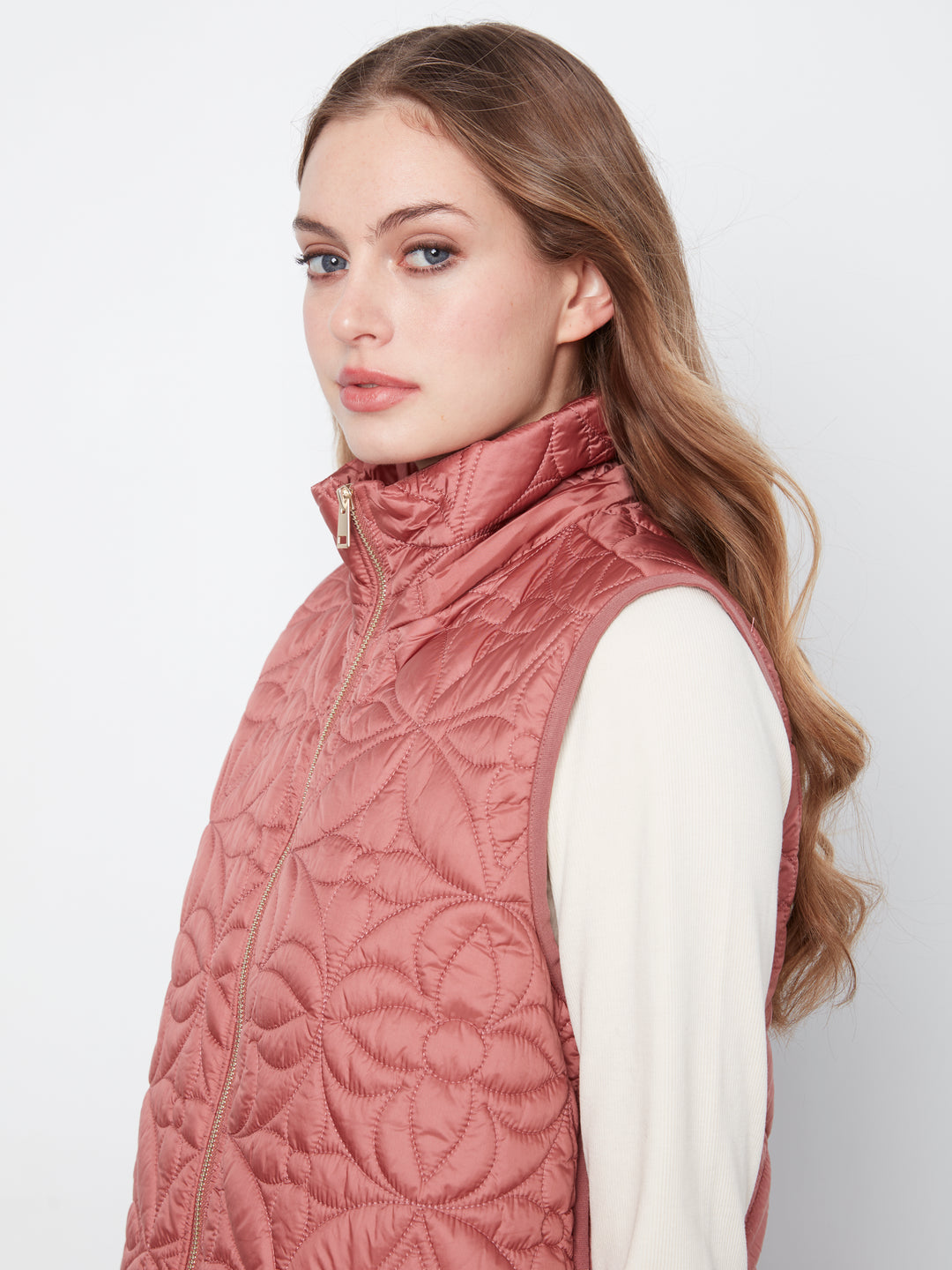 A woman wearing a sleeveless quilted vest in a dusty rose pink color, layered over a white long-sleeve top, photographed against a light gray studio background.