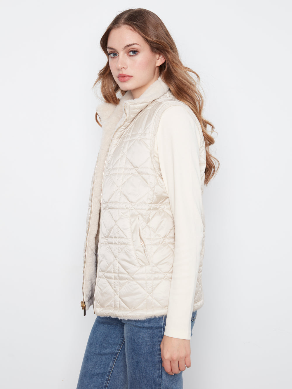 A woman wearing a cream-colored quilted sleeveless puffer vest over a matching cream long-sleeve top and blue jeans, photographed against a plain white studio background.