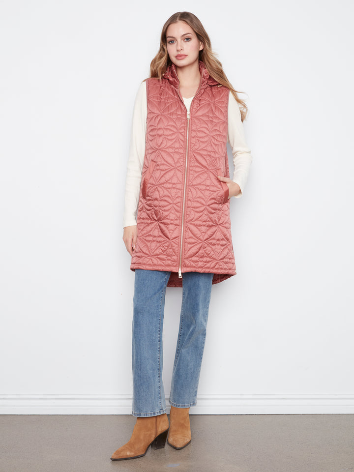 A woman models a dusty rose pink quilted sleeveless puffer vest with a front zipper, worn over a cream long-sleeve top, blue jeans, and tan suede ankle boots against a white studio background.