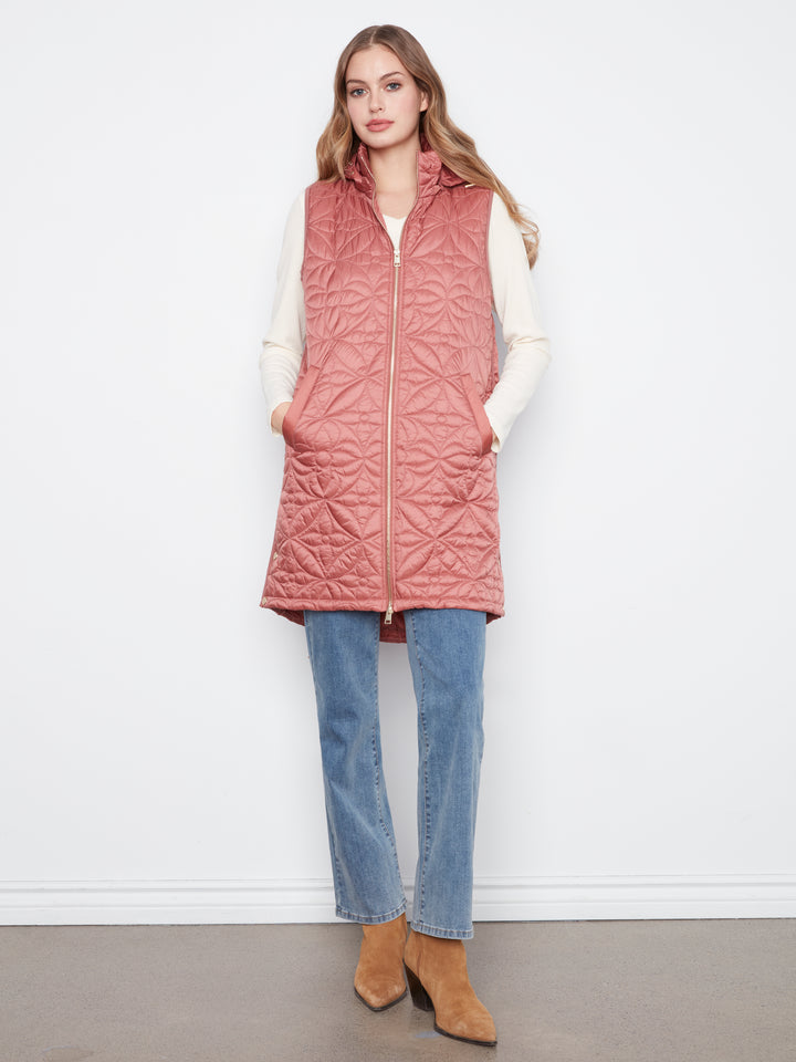 A woman modeling a dusty rose quilted zip-front vest over a cream long-sleeve top, blue jeans, and tan ankle boots, standing against a plain white wall.