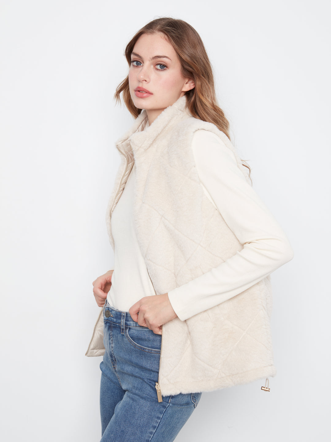 A woman wearing a cream-colored quilted sleeveless vest over a matching cream long-sleeve top and blue jeans, photographed against a plain white studio background.