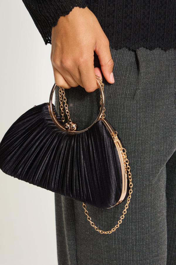 A close-up of a person holding a pleated black velvet clutch bag with gold-tone hardware and chain straps, paired with dark gray trousers.