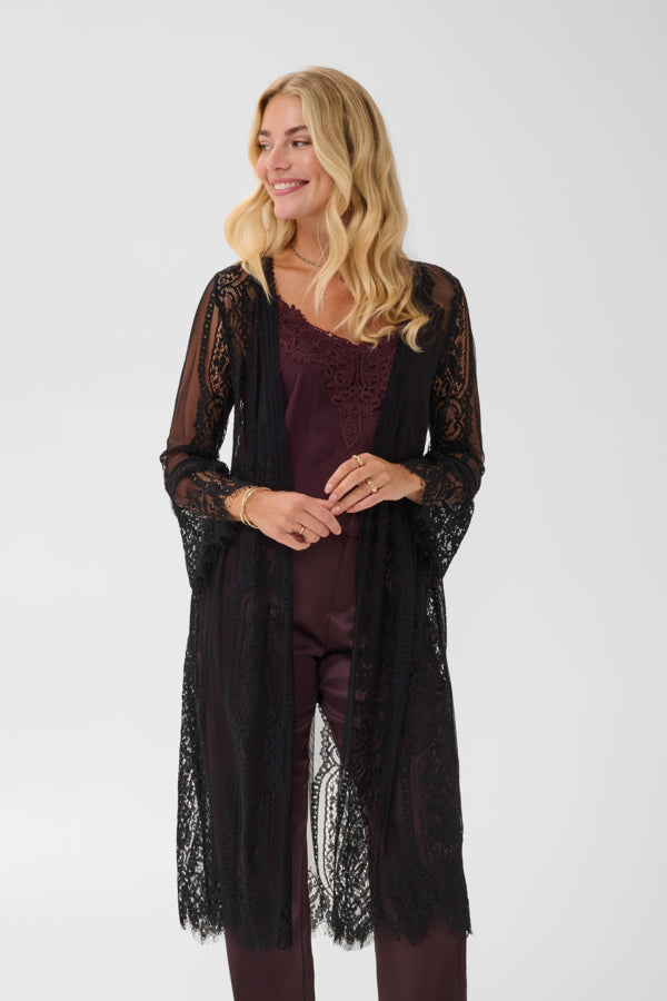 A woman modeling a long black sheer lace cardigan over a burgundy top and matching pants, photographed against a plain light gray studio background.