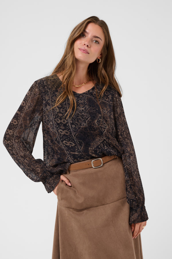 A woman modeling a sheer black long-sleeve blouse with an intricate brown paisley pattern, paired with a brown belt and brown skirt, against a light gray studio background.