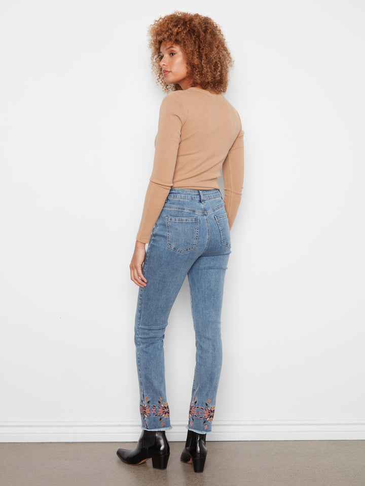 A woman with curly brown hair is shown from the back wearing light-wash blue jeans with floral embroidery at the cuffs, a beige long-sleeve top, and black ankle boots, standing against a white wall.