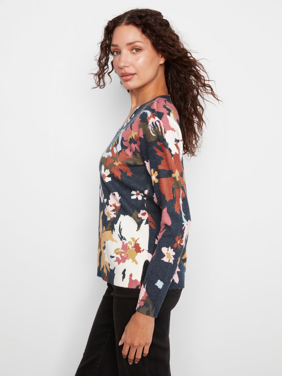 A woman wearing a long-sleeve knit sweater with a multicolor floral and abstract pattern on a dark navy base, paired with black pants, photographed against a light gray studio background.