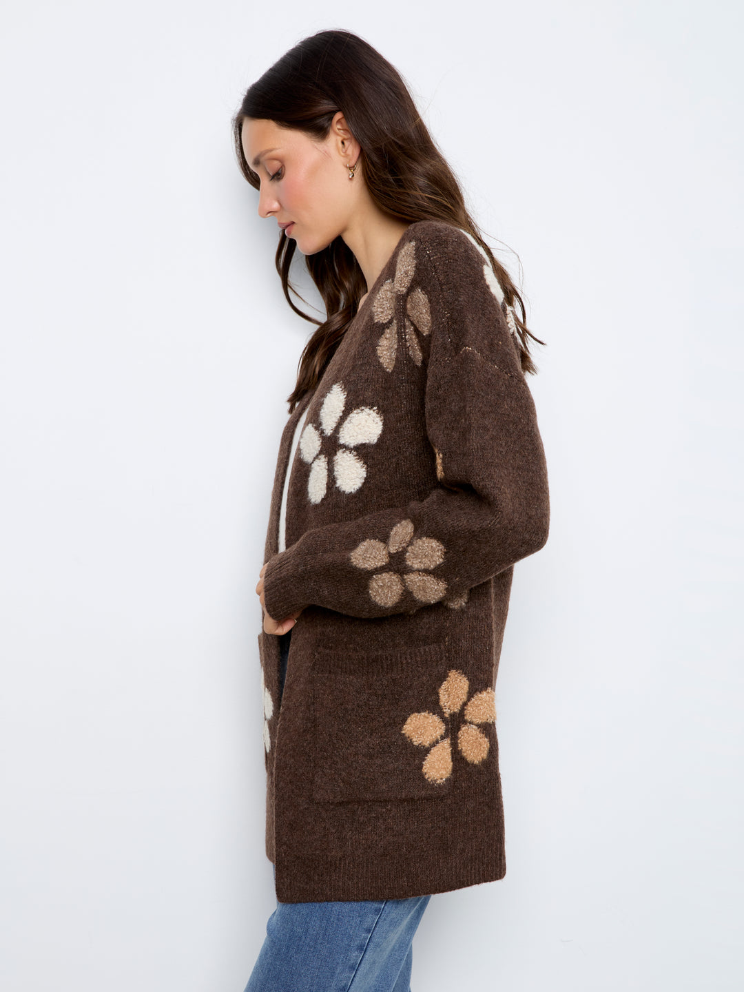 A woman wearing a brown knit cardigan with large beige and cream floral patterns, paired with blue jeans, photographed against a plain white studio background.