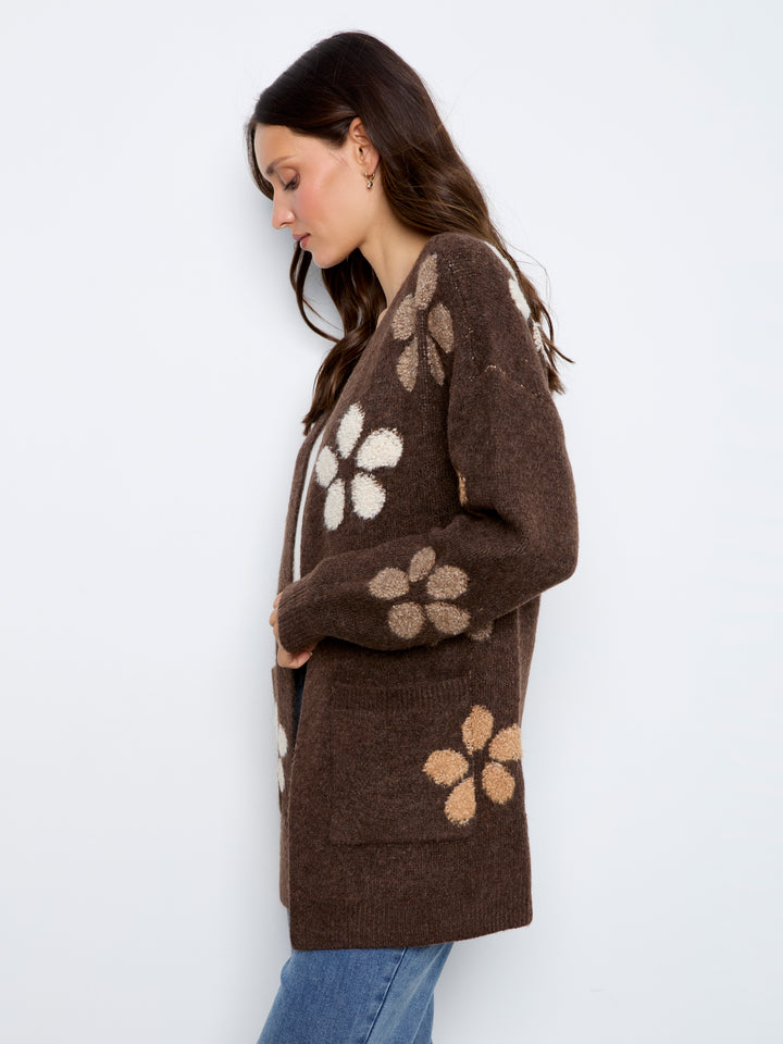A woman wearing a brown knit cardigan with large beige and cream floral patterns, paired with blue jeans, photographed against a plain white studio background.