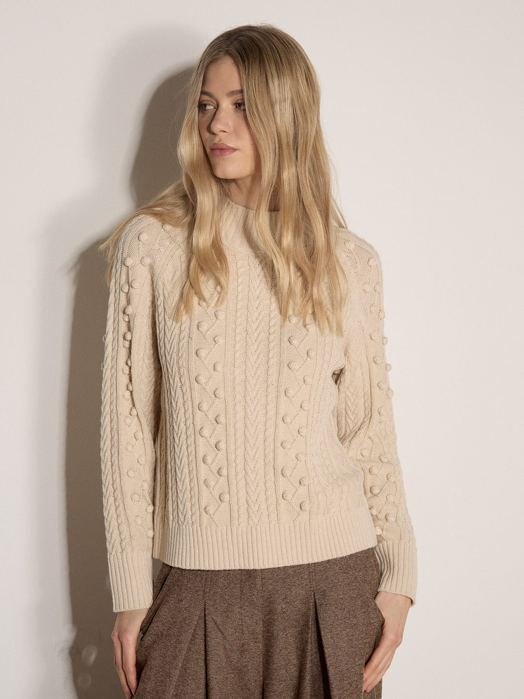 A woman wearing a cream-colored cable-knit sweater with textured pom-pom details on the sleeves, paired with brown pleated trousers, standing against a plain off-white wall.