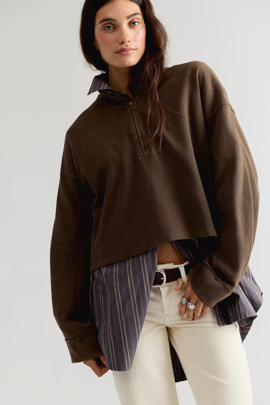 A model wearing a brown cropped quarter-zip pullover sweater, styled with a striped shirt and white pants against a light gray studio background.