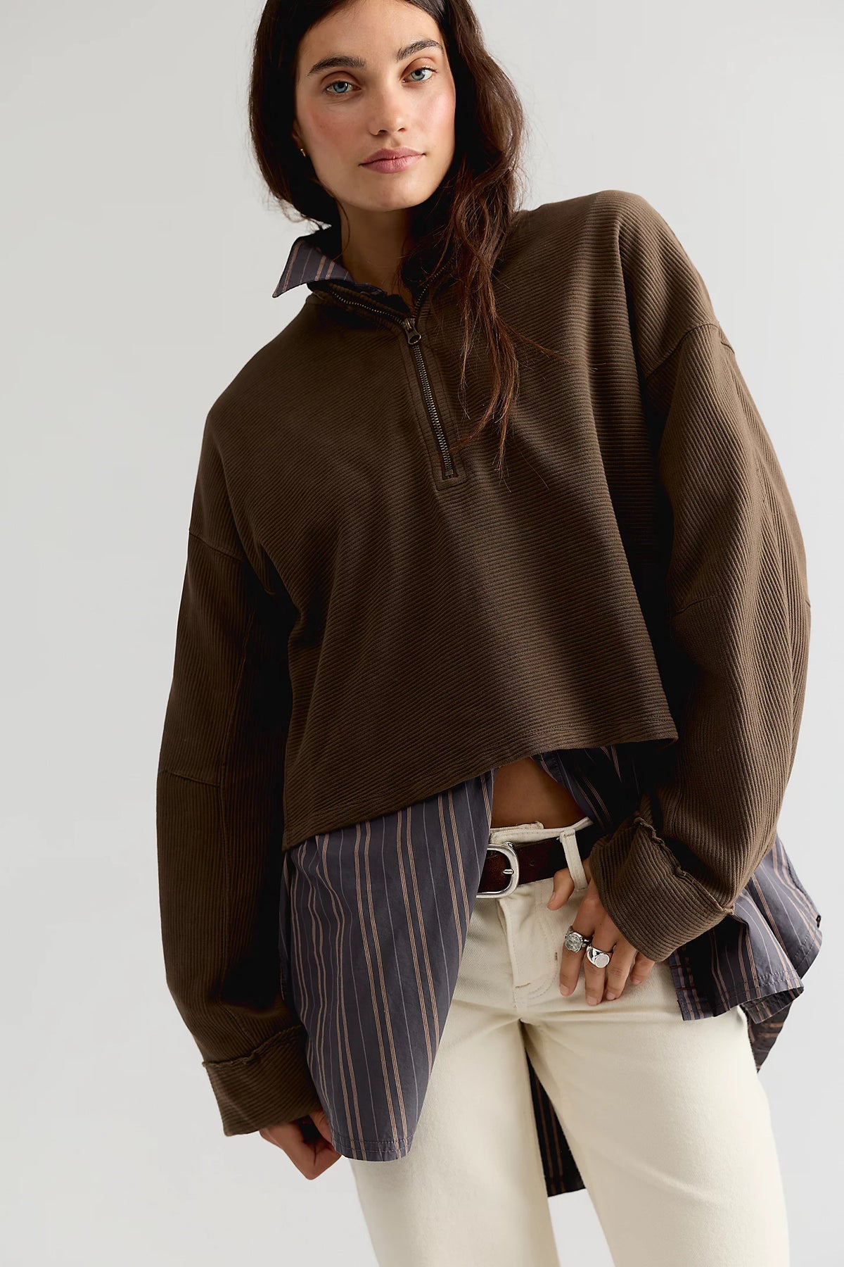 A model wearing a brown cropped quarter-zip pullover sweater, styled with a striped shirt and white pants against a light gray studio background.