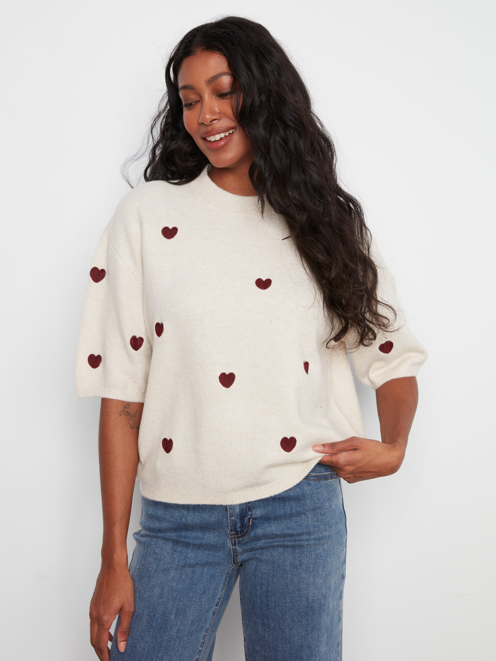 Woman wearing a white sweater with red heart patterns and blue jeans on a plain background