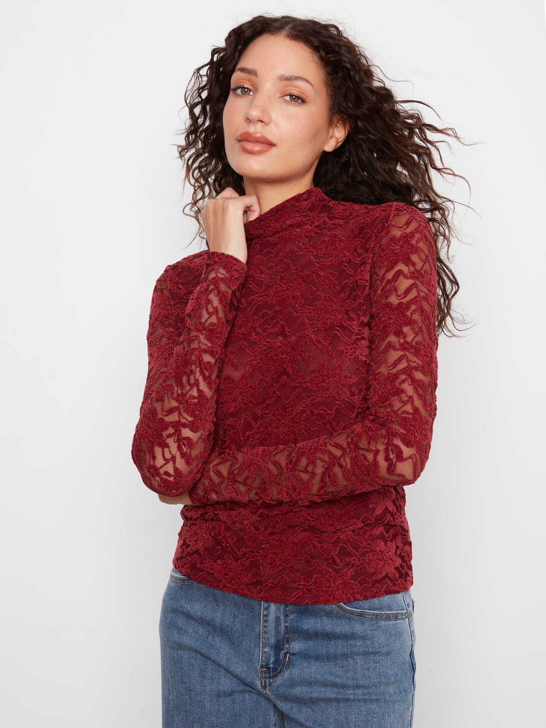 A woman with long, curly dark hair wearing a textured, long-sleeve burgundy lace mock-neck top and blue jeans, posing against a plain white studio background.