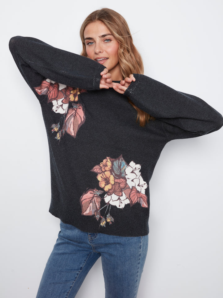 A woman wearing a dark charcoal gray knit sweater with a large floral print in pink, white, and yellow, paired with blue jeans, posing against a light gray background.