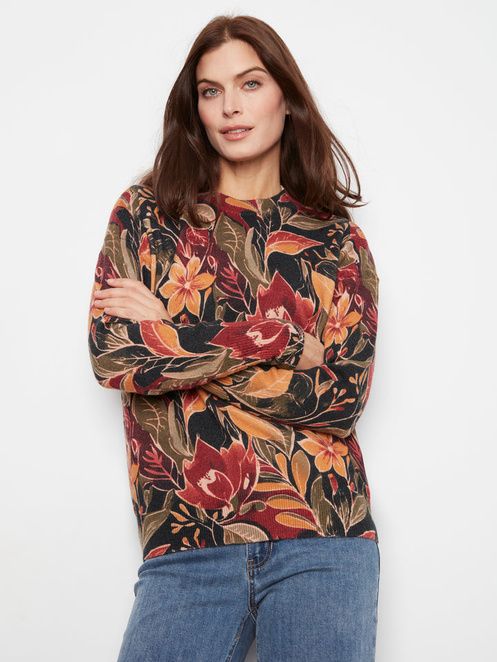 A woman wearing a long-sleeve crewneck sweatshirt with a dense tropical floral and leaf pattern in red, orange, brown, and beige tones, paired with blue jeans, against a plain white studio background.