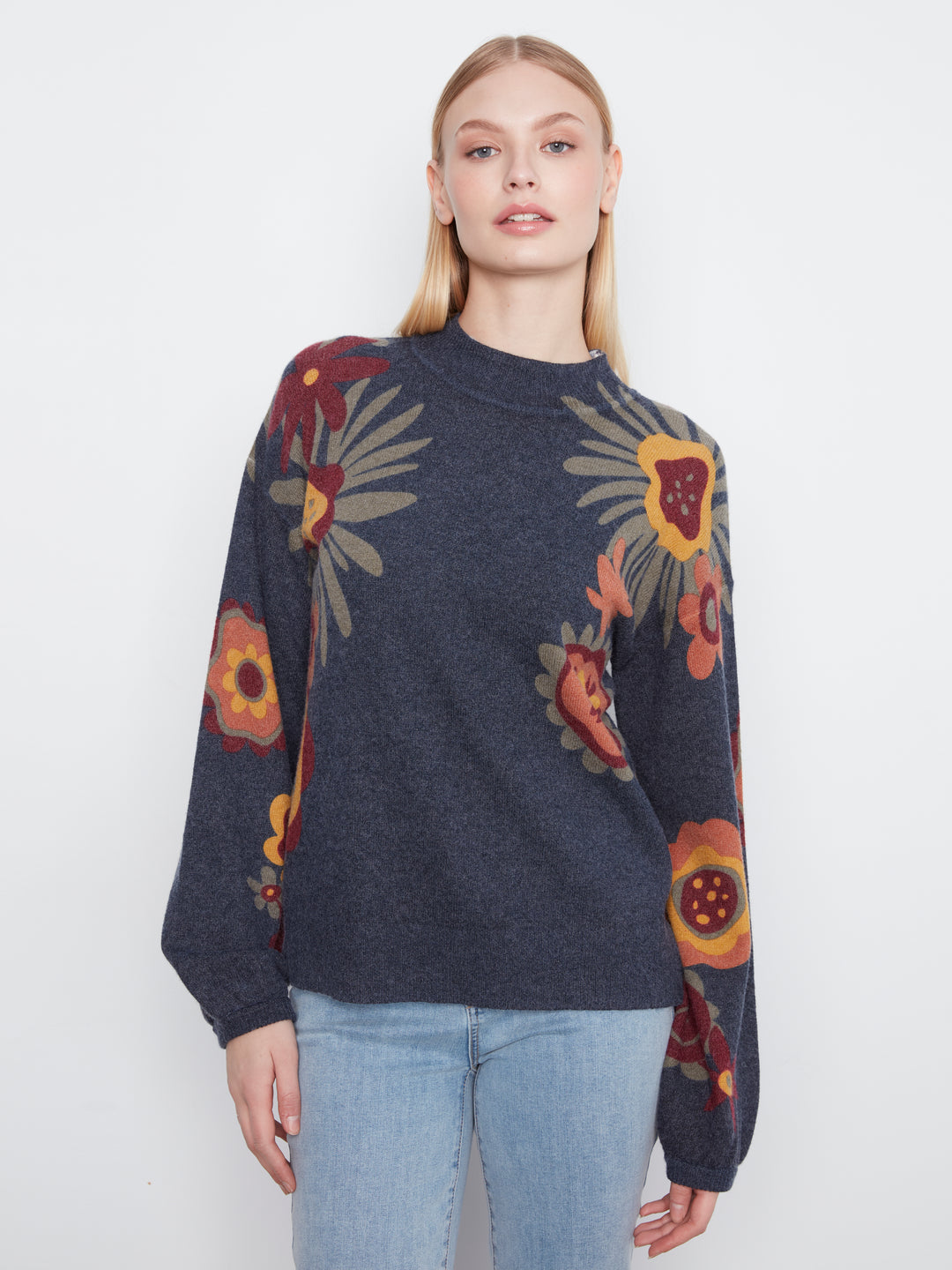 A woman modeling a dark navy blue knit sweater with a colorful floral pattern in yellow, orange, and red, paired with light blue jeans against a plain white background.