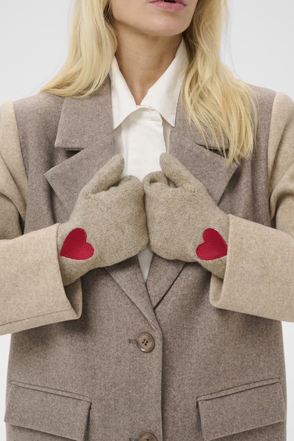 A close-up of a woman wearing beige knit winter gloves with red heart patches, paired with a taupe wool coat and white collared shirt.