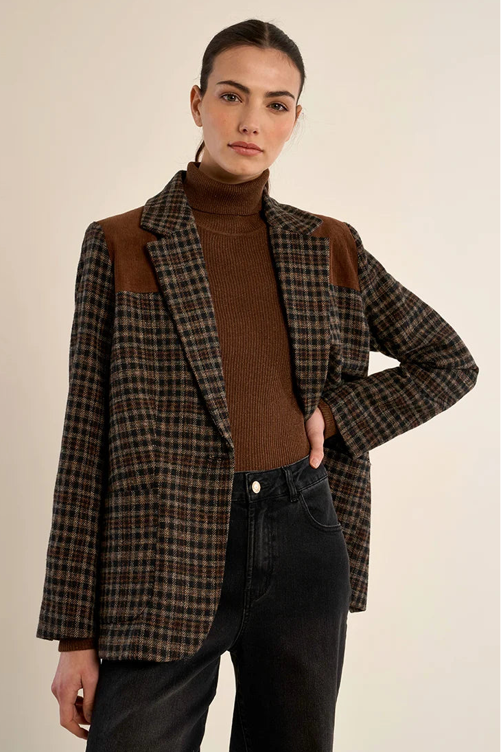 A model wearing a brown and black plaid blazer with brown suede shoulder patches, styled over a brown turtleneck sweater and black jeans, against a plain beige background.