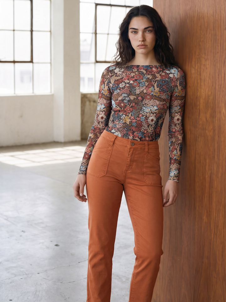 A woman stands against a wooden wall in a bright industrial loft, wearing a fitted long-sleeve floral top and rust-orange cropped pants.