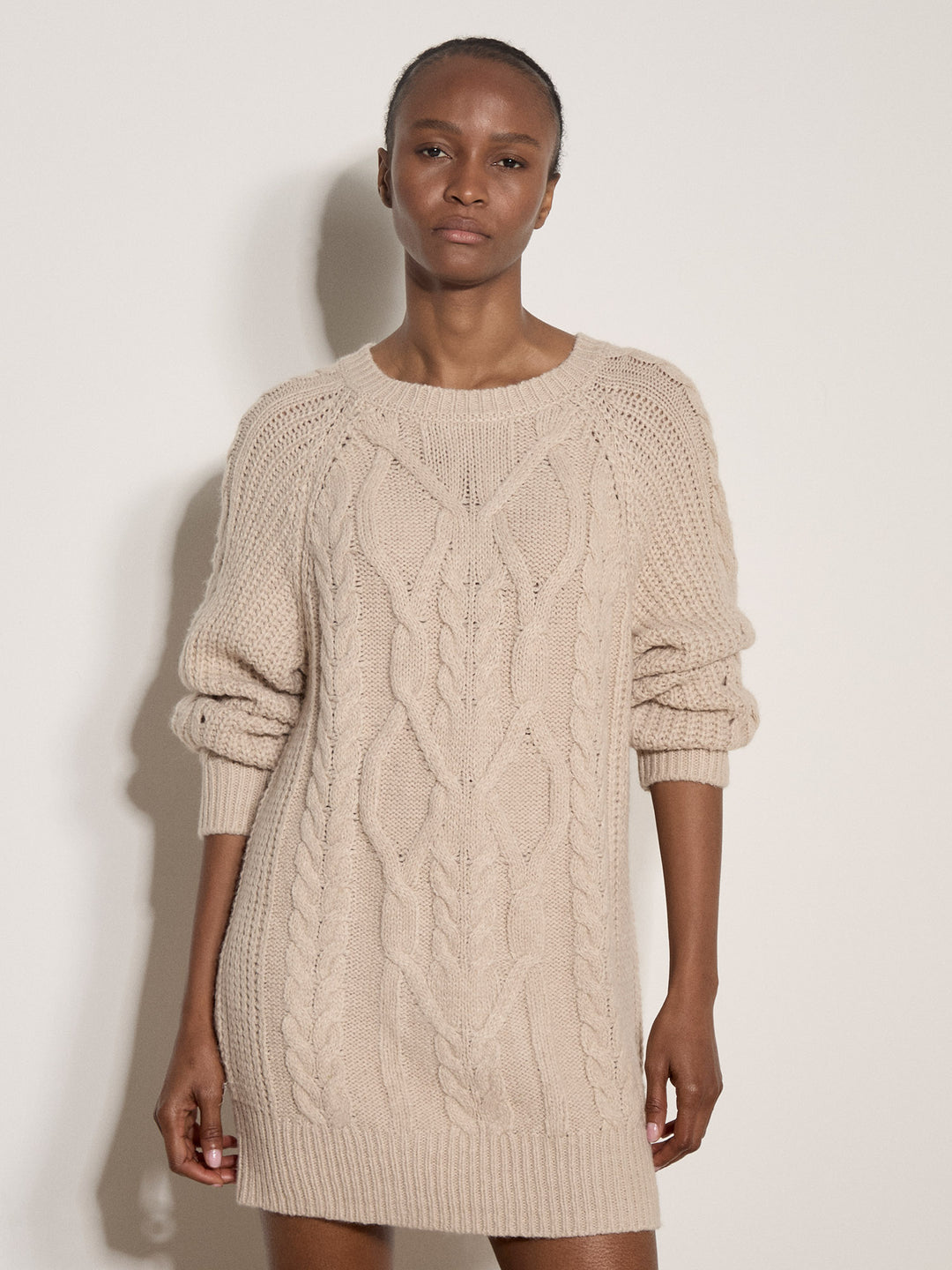 A model wearing a chunky cable-knit sweater dress in a light beige color, standing against a plain off-white wall.