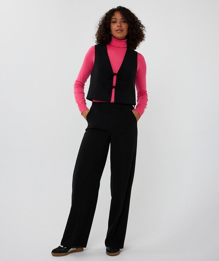 Person wearing a pink turtleneck, black vest, and black pants on a white background