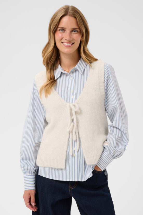 A woman modeling a cream-colored knit vest over a blue and white striped button-down shirt, paired with dark blue jeans, against a light gray studio background.