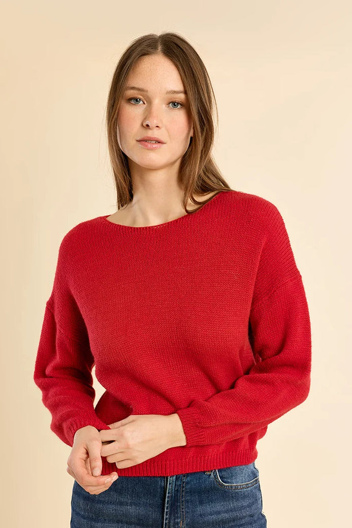 A woman wearing a bright red knit sweater with a wide boat neckline and blue jeans, standing against a solid beige studio background.