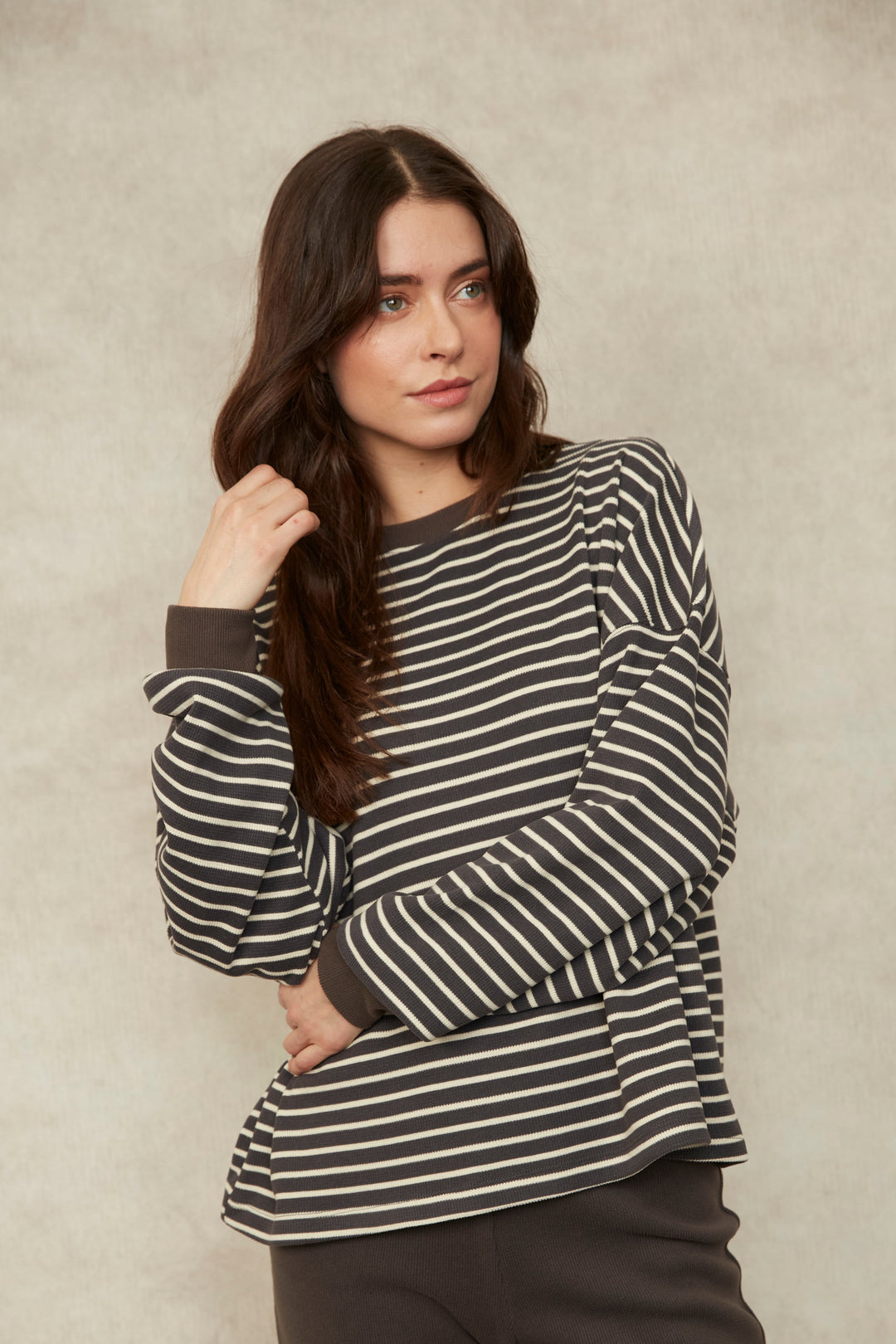A woman modeling a black and white horizontally striped long-sleeve top with dark brown pants, standing against a textured beige studio backdrop.