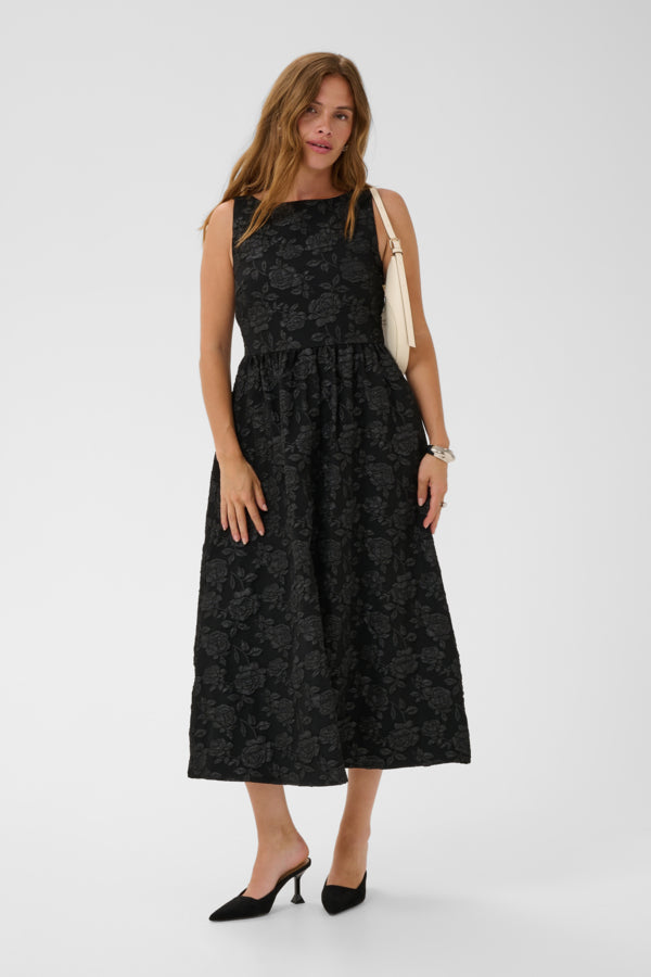 A woman modeling a sleeveless black midi dress with a subtle tonal floral jacquard pattern, styled with a cream shoulder bag and black pointed-toe mule heels against a light gray studio background.