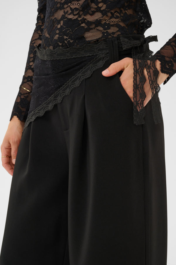 A close-up of a model wearing black wide-leg trousers with a matching black lace wrap belt and a black lace long-sleeve top, with one hand in the trouser pocket.