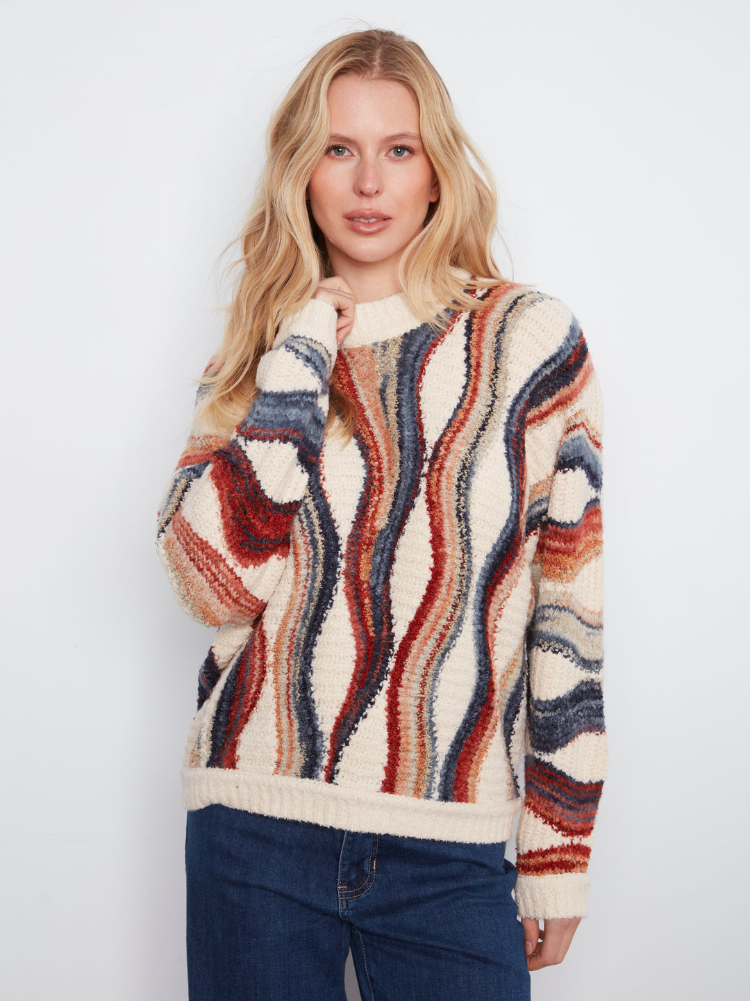 A woman wearing a cream-colored knit sweater with vertical wavy stripes in rust red, navy blue, and brown, paired with dark blue jeans, against a plain light gray background.
