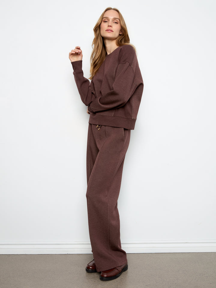 A woman modeling a matching brown lounge set consisting of a long-sleeve sweatshirt and wide-leg trousers, standing against a plain white wall.