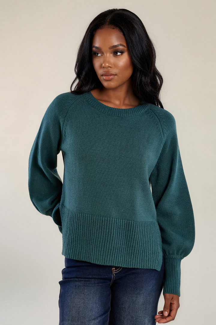 A woman modeling a dark teal green knit sweater with a crew neck and ribbed hem, paired with blue jeans, against a plain beige background.