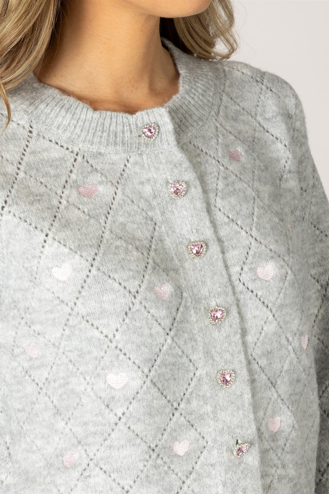 Close-up of a light gray knit cardigan sweater with a diamond pattern, pink heart details, and rhinestone-embellished buttons, worn by a model.