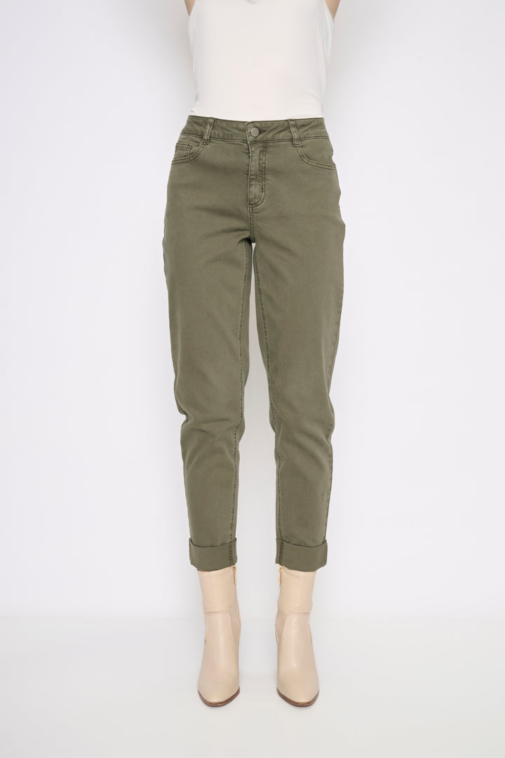 A model is wearing a pair of olive green cropped jeans with rolled cuffs, paired with a white sleeveless top and beige ankle boots, shot against a plain white studio background.
