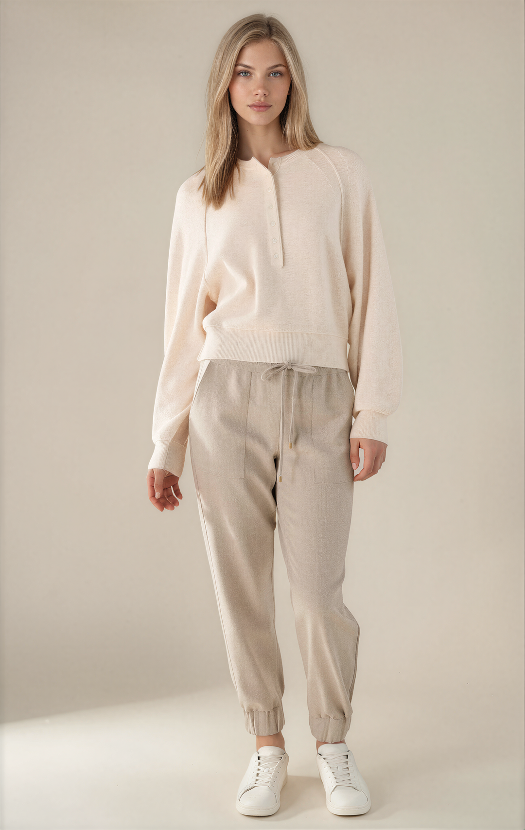 A woman modeling a light cream knit long-sleeve top and beige jogger pants with white sneakers, standing against a plain beige studio background.