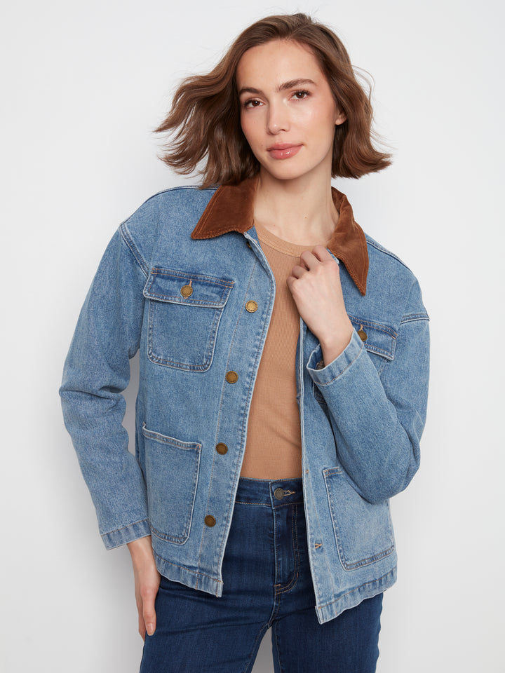 A woman wearing a light blue denim jacket with a brown suede-like collar, layered over a tan top and dark blue jeans, photographed against a plain light gray studio background.