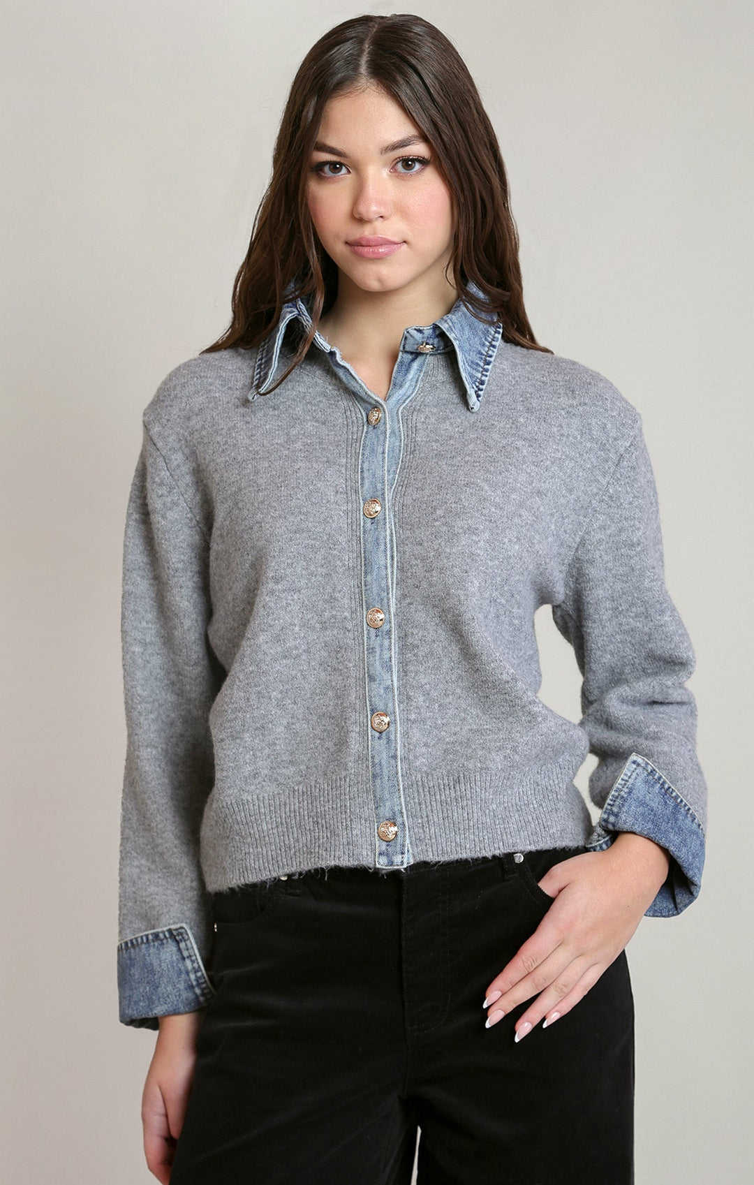 Woman wearing a gray sweater over a denim shirt with black pants on a plain background