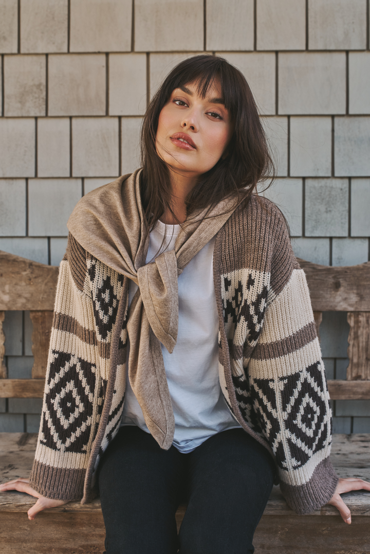 A woman sits on a rustic wooden bench wearing a beige and brown knit cardigan with a geometric diamond pattern and a matching beige knit wrap tied around her neck, over a white top and black pants, set against a light gray shingled wall.