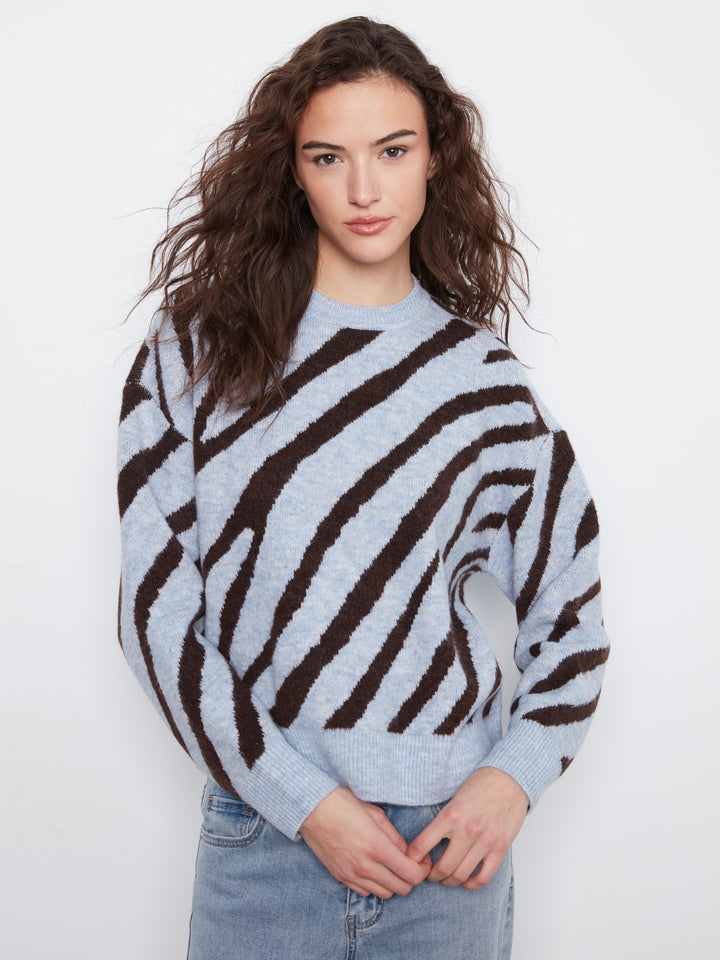 A woman wearing a light gray and dark brown zebra-striped knit sweater with light-wash blue jeans, standing against a plain white studio background.