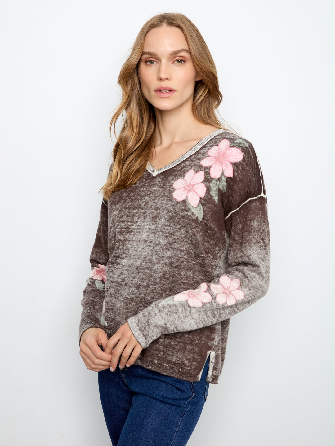 A woman wearing a brown marled knit sweater with pink floral embroidery on the shoulders and sleeves, paired with blue jeans, photographed against a plain white studio background.