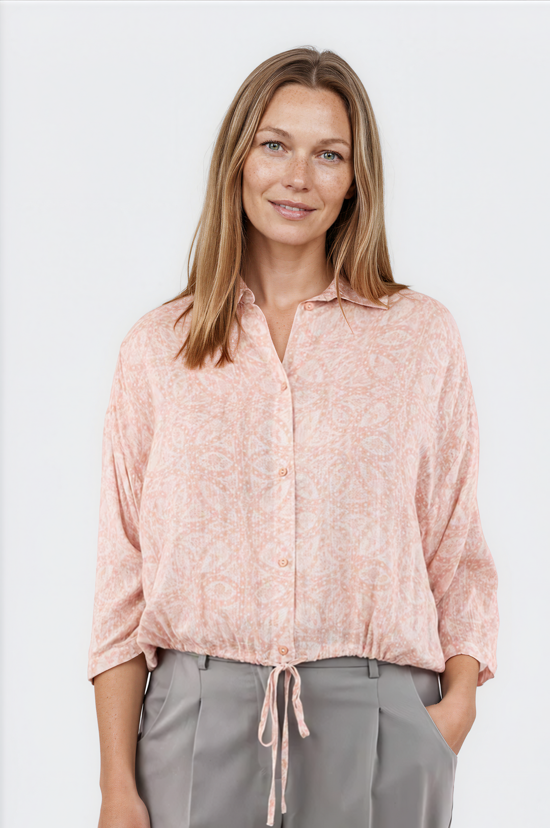 Woman wearing a light pink blouse with a subtle pattern and gray pants on a white background