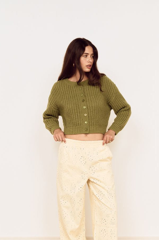 A woman modeling a cropped olive green knit cardigan with buttons and cream wide-leg eyelet trousers, standing on a light wood herringbone floor against a white wall.