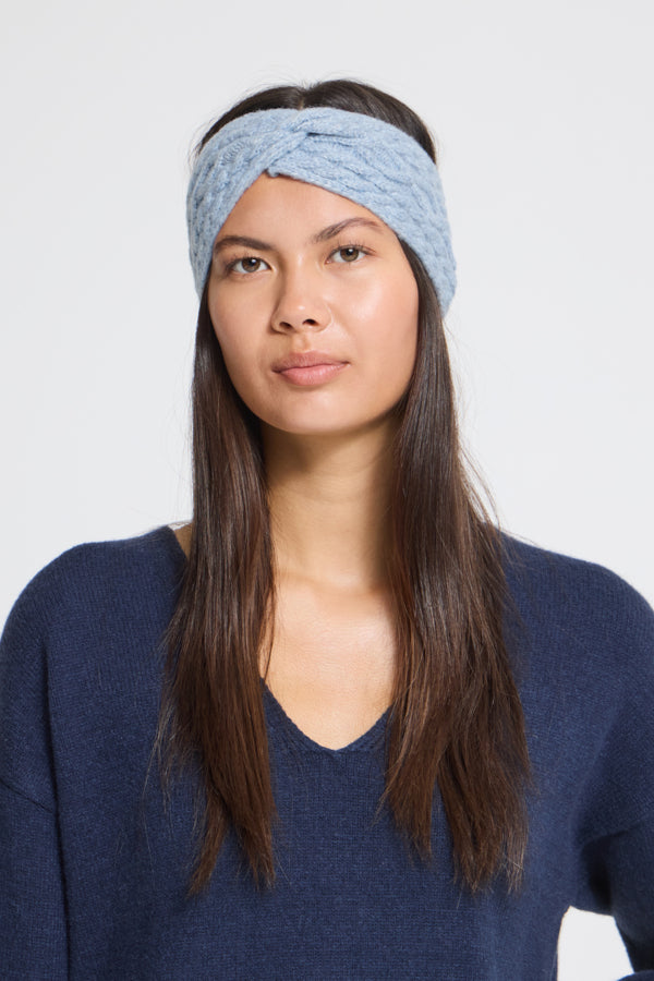 A woman wearing a light blue twisted knit headband and a navy blue V-neck sweater, photographed against a plain light gray studio background.