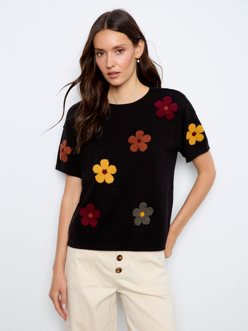 A woman modeling a black short-sleeve knit top with colorful 3D flower appliqués in yellow, red, orange, and gray, paired with cream pants against a light gray studio background.