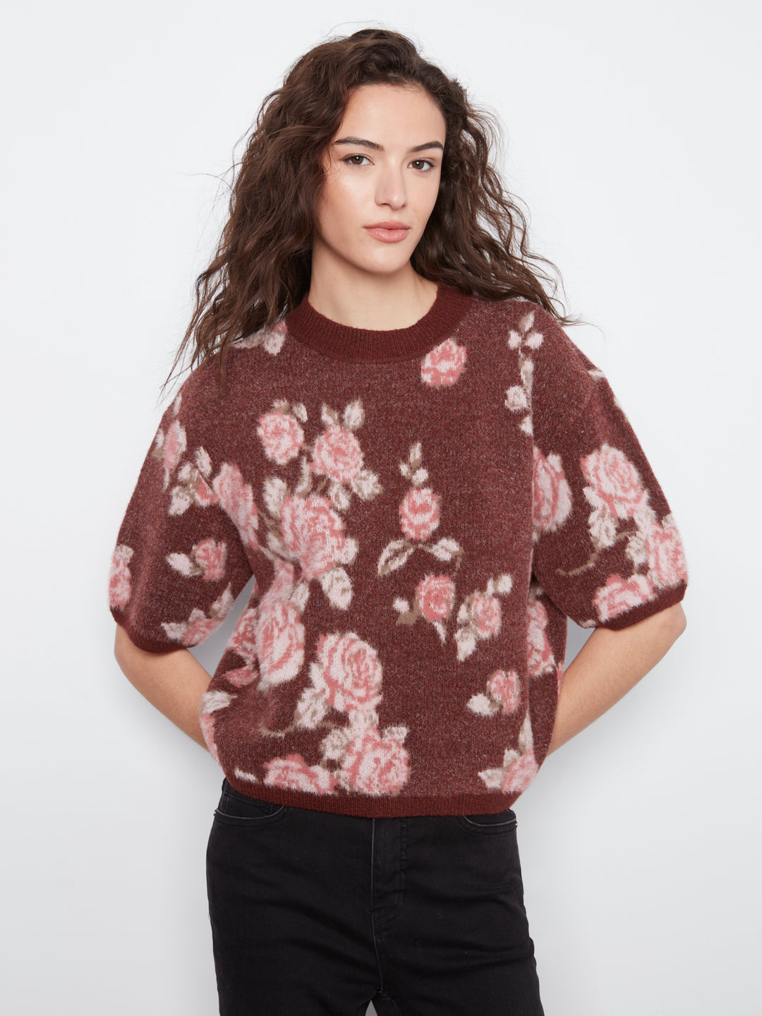 A woman wearing a short-sleeve crewneck sweater with a pink and white floral pattern on a dark brown background, paired with black pants, against a light gray studio background.