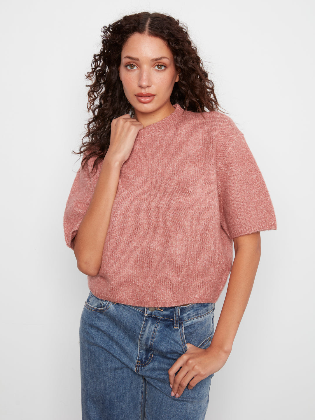 A woman with curly dark hair wearing a dusty rose short-sleeve knit top and blue jeans, standing against a light gray studio background.