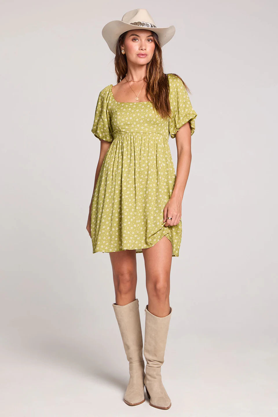 Woman wearing a green dress with beige boots and a white hat on a plain background