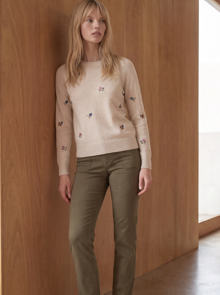 A woman stands barefoot against a warm wood-paneled wall, wearing a light beige crewneck sweater with small embroidered details and olive green trousers.